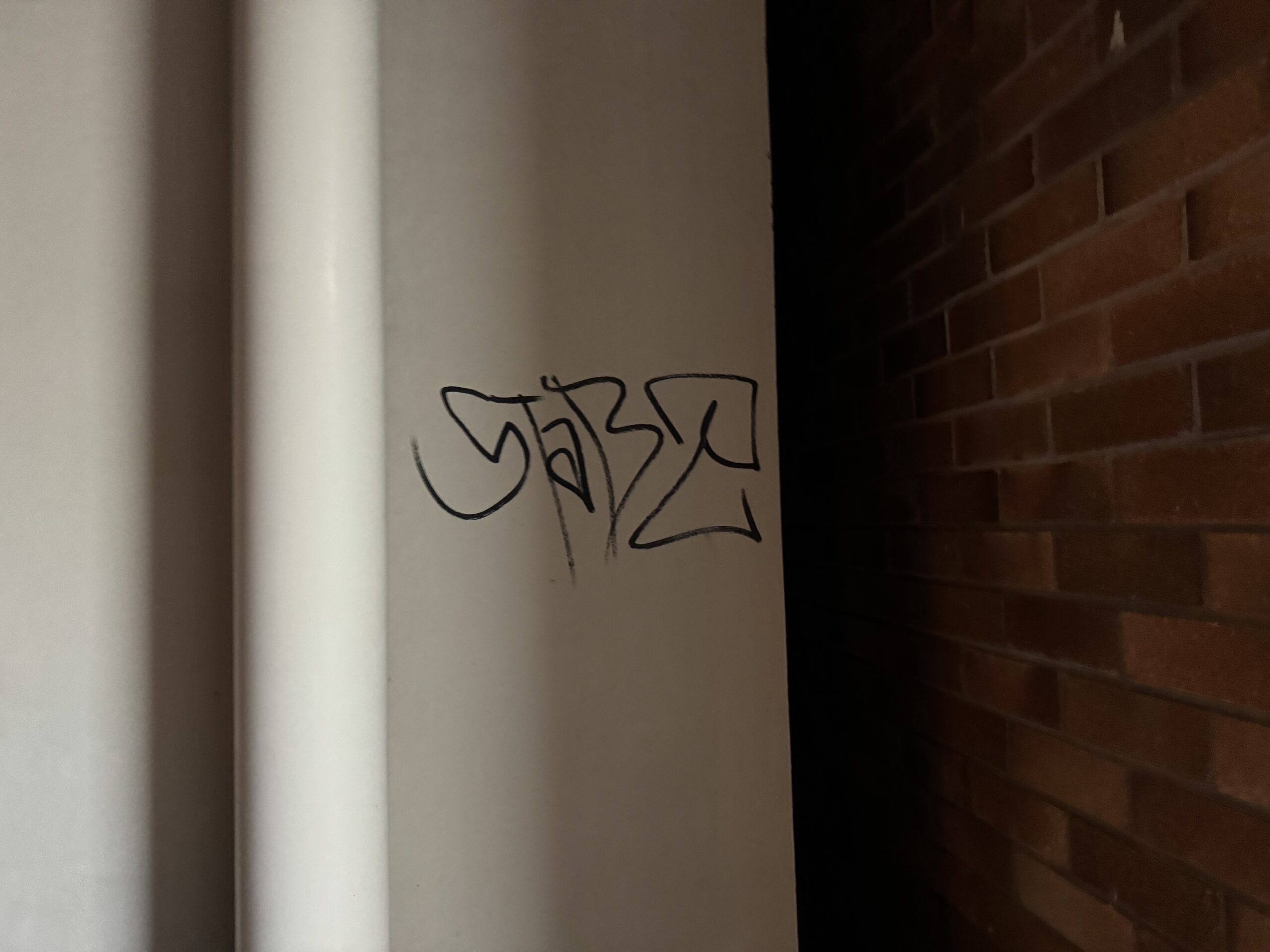 Black graffiti tag on a cream painted wall beside a downpipe and brickwork